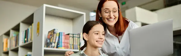 stock image A redheaded woman holds a laptop in front of a bookshelf while teaching a teenage girl, redefining modern education.