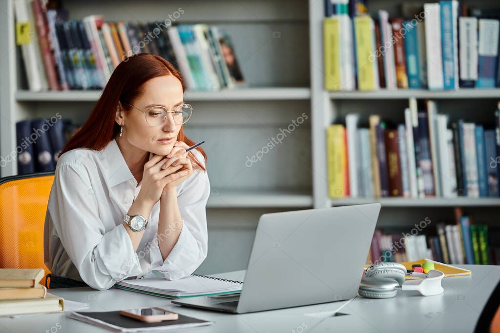 A redheaded female tutor is teaching an online lesson using a laptop at a desk.