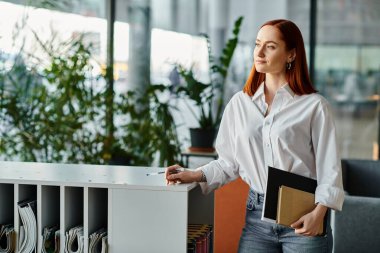 Kızıl saçlı kadın elinde defter ve kitapla modern eğitim sahnesiyle duruyor..
