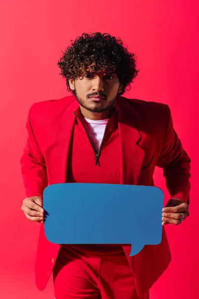 stock image Handsome young Indian man in vibrant red suit holds a speech bubble against a vivid backdrop.