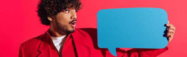 Young Indian man in vibrant red jacket holding a blue speech bubble.