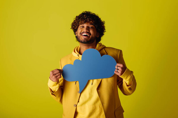 Handsome Indian man in yellow jacket holds blue speech bubble on vivid backdrop.