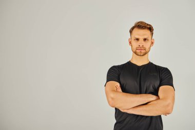 Siyah tişörtlü genç atletik adam güçlü bir duruş sergiliyor. Gri stüdyo arkaplanına karşı güvenli bir jest yaparak kollarını kavuşturuyor..