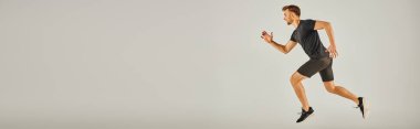 Aktif giyinmiş genç bir atletik, gri bir arka planda, stüdyoda enerjik bir şekilde koşuyor..