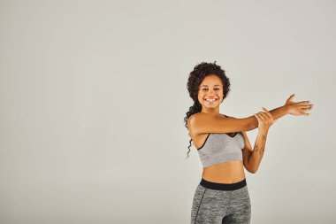 Aktif giyinmiş genç bir Afrikalı Amerikalı kadın gri bir arka plan önünde nazikçe kollarını açıyor..