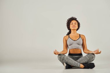Aktif giyinmiş genç bir Afrikalı Amerikalı kadın gri bir arka planda, stüdyo ortamında huzur içinde meditasyon yapıyor..