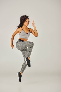 Genç, formda, spor sutyenli ve taytlı bir Afro-Amerikan kadın gri bir stüdyonun arka planında güçlü bir şekilde zıplıyor..