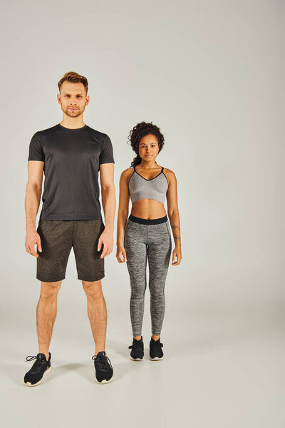 A young interracial sport couple in active wear stands side by side on a grey studio background.