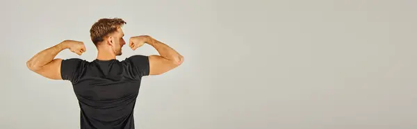 stock image A young athletic man in active wear flexing his muscles in front of a gray background in a studio.