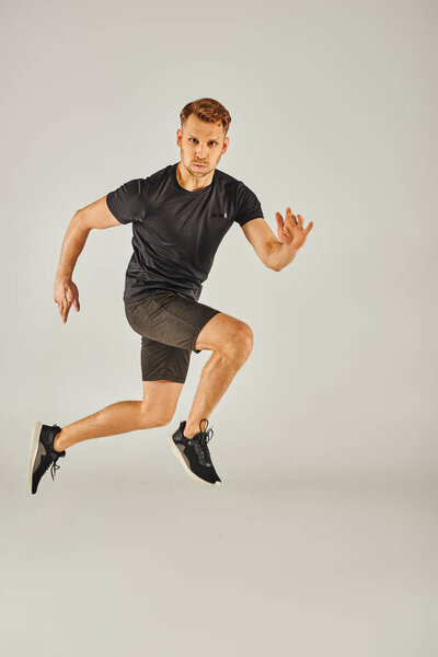 A young athletic man in a black t-shirt is energetically jumping in a studio with a grey background.