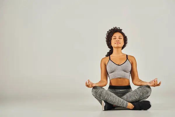 stock image A young African American woman in activewear meditates peacefully on a gray background in a studio setting.