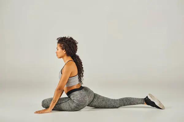 A young African American woman in active wear gracefully stretches in a squat position on a grey background.