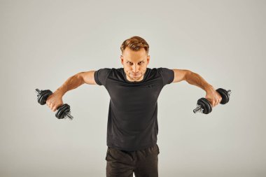 Aktif giyinen genç bir sporcunun gri arka planı olan bir stüdyoda iki dambılı var..