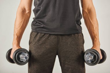 Young sportsman in active wear holding two dumbbells in his hands, exercising in a studio with a grey background. clipart