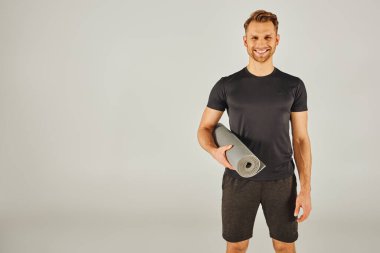 Aktif giyinmiş genç bir adam gri bir arka planın önünde yoga minderi tutuyor..
