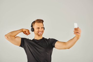 Aktif giyinen genç bir sporcu gri arka planı olan bir stüdyoda telefonuyla selfie çekiyor..