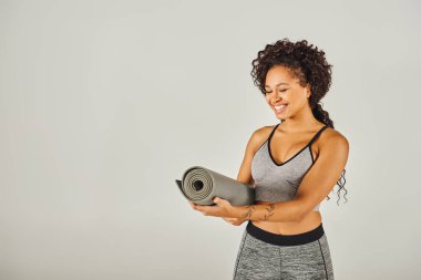 Spor kıyafetli kıvırcık Afrikalı Amerikalı bir sporcu gri arka planı olan bir stüdyoda yoga minderi tutuyor..
