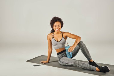 Curly African American sportswoman in active wear sitting on yoga mat, holding water bottle, with grey studio background. clipart