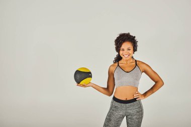 Aktif giyinen kıvırcık saçlı Afrikalı Amerikalı sporcu, stüdyo ortamında siyah ve sarı bir top tutuyor..
