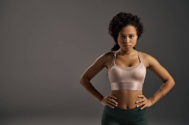 Spor sutyenli kıvırcık Afrikalı Amerikalı sporcu, gri arka planlı bir stüdyoda güven içinde poz veriyor..