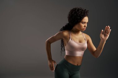 Spor sutyen ve taytlı kıvırcık Afrikalı Amerikalı sporcu gri bir arka planla fotoğraf çekiminde poz veriyor..