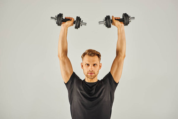 Молодой спортсмен в активной одежде поднимает гантели в студии на сером фоне.