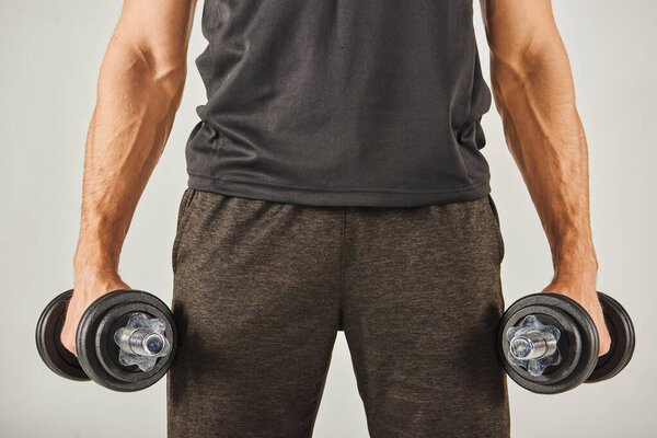 Young sportsman in active wear holding two dumbbells in his hands, exercising in a studio with a grey background.