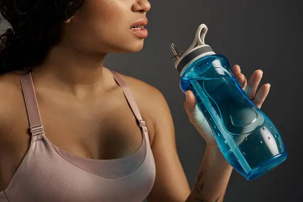 stock image African American sportswoman in active wear drinking from a blue water bottle in a studio with a grey background.