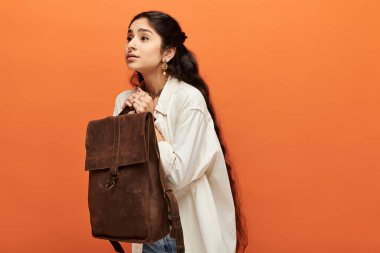 Genç Hintli bir kadın kahverengi bir sırt çantasını canlı turuncu bir arka plana yaslar..