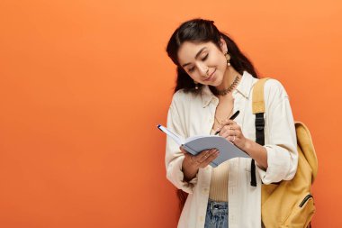 Young indian woman actively writing in notebook against bright orange background. clipart