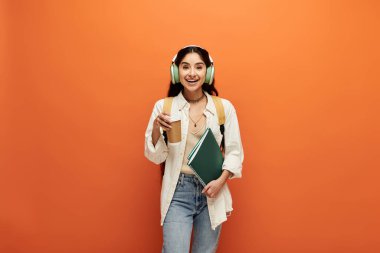 Young indian woman wearing headphones holds book against orange backdrop. clipart