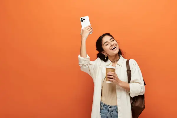 stock image indian woman capturing moment with phone on vibrant orange backdrop.