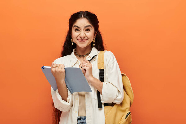 Young indian woman energetically holds a notebook against a vibrant orange background.