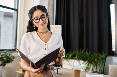 A dynamic indian woman with glasses handling a folder in a professional office setting. clipart