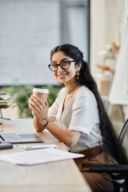 Gözlüklü genç bir Hintli kadın masasında bir fincan kahve içiyor..