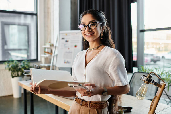 A professional indian woman with glasses holding a folder in an office setting.