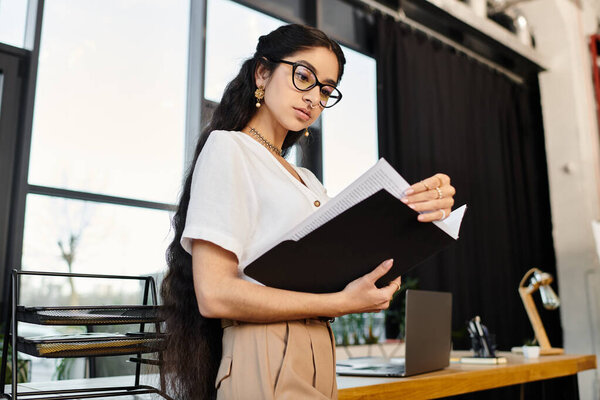 A stylish indian woman in glasses confidently holds a folder in a sleek office setting.