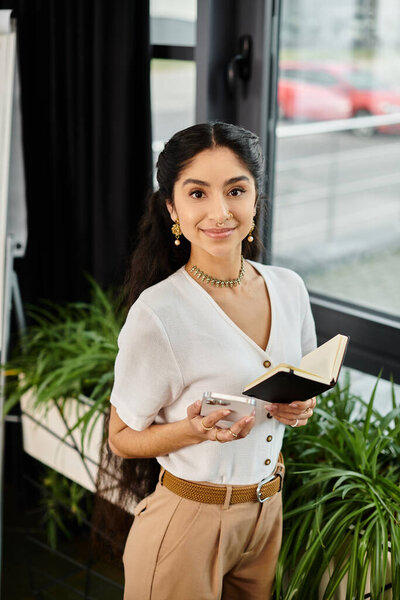 Young indian woman holding a book in front of a lush plant, connecting with nature.