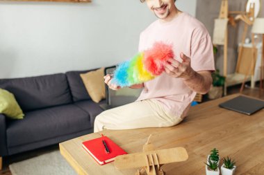 A man in cozy homewear sits on a table, holding a vibrant duster. clipart