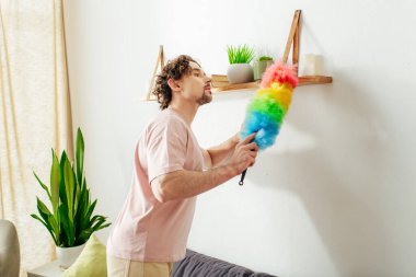 Handsome man in cozy homewear holding a vibrant rainbow duster. clipart