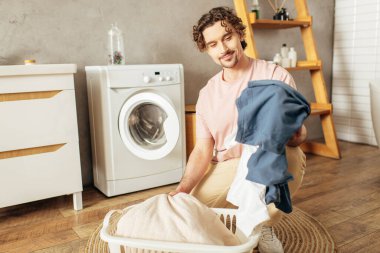 A man in cozy homewear holds a laundry bag in front of a washing machine. clipart