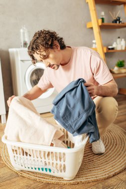Handsome man in cozy homewear neatly arranging clothes in laundry basket. clipart
