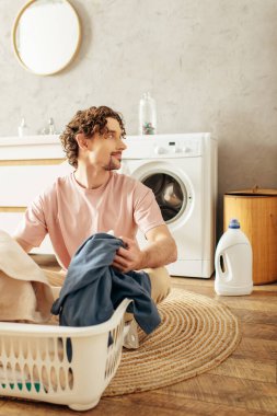A handsome man in cozy homewear sits beside a laundry basket. clipart