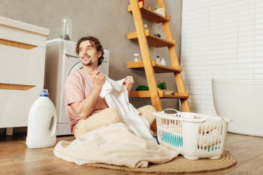 Handsome man in homewear sitting on floor sorting laundry. clipart