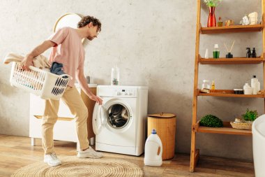 Çamaşır sepetini çamaşır makinesiyle tutan rahat ev elbiseli yakışıklı bir adam..
