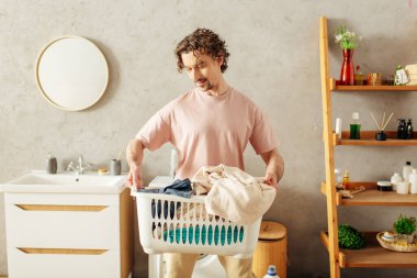 Handsome man in cozy homewear holds laundry basket in bathroom. clipart