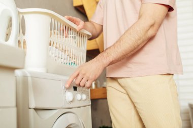 Handsome man in cozy homewear putting laundry basket on washing machine. clipart