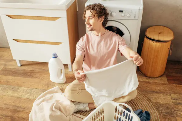 stock image A handsome man in cozy homewear sitting beside a washing machine.