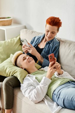 İki kadın, kısa saçlı lezbiyen bir çift, kendilerini telefonlarına kaptırmış bir kanepede oturuyorlar..