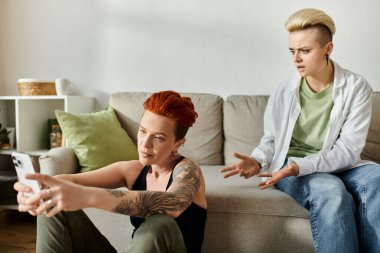 Two women with short hair engaged in conversation about online cheating while sitting on a sofa in a living room. clipart